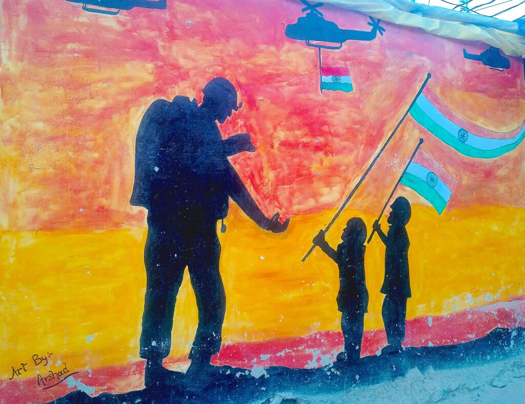 With human figures painted as dark silhouettes, a large mural depicts a soldier wearing a helmet and large backpack reaching toward two children holding flags with orange, white, and green horizontal stripes. They stand against a bright yellow, orange, and red background with three black helicopters—one displaying the same flag—flying overhead.