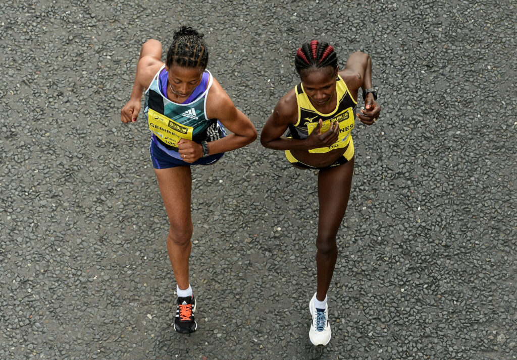 Taken from above, a photograph shows two people running. One wears a blue-and-green colored tank top, shorts, and sneakers, while the other wears a similar outfit in yellow and black.