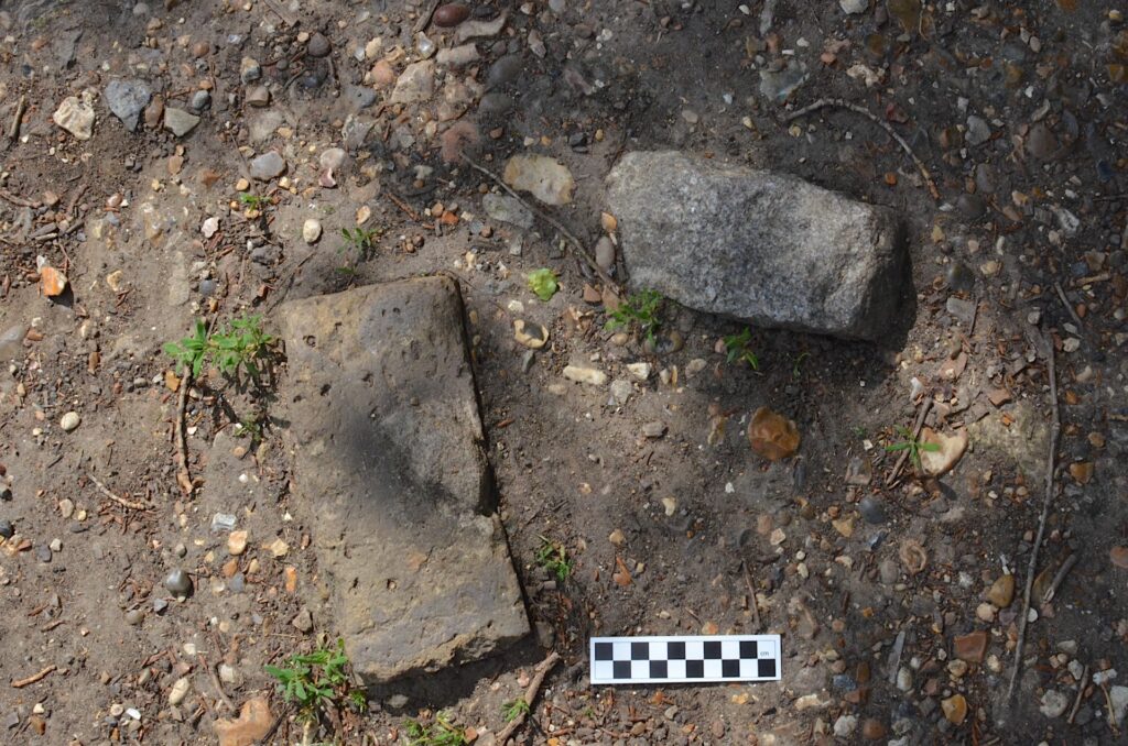 Two large stones sit on a ground of dirt and much smaller pebbles. A checkered black-and-white ruler lies next to them.