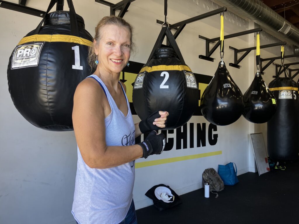 A smiling person with their hair in a ponytail wears teal earrings, a gray sleeveless top, and black open-finger gloves while standing in front of several punching bags.