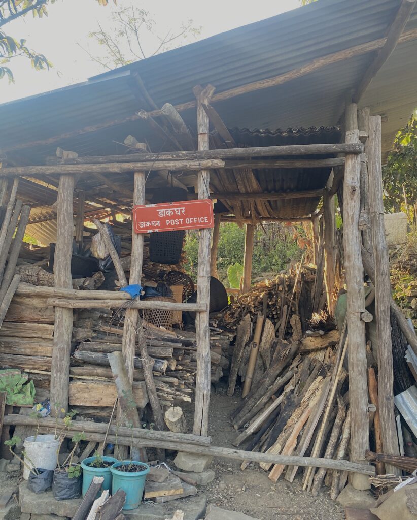 A structure of wooden beams and a ridged tin roof has a red sign posted on its front. Blue pots and several piles of wood sit inside the structure.
