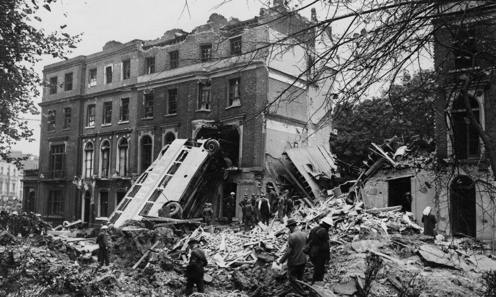 A black-and-white photograph shows a large bus leaning on its side against a four-story brick building. Several people in suits are standing around in a pile of rubble surrounding the wreck.