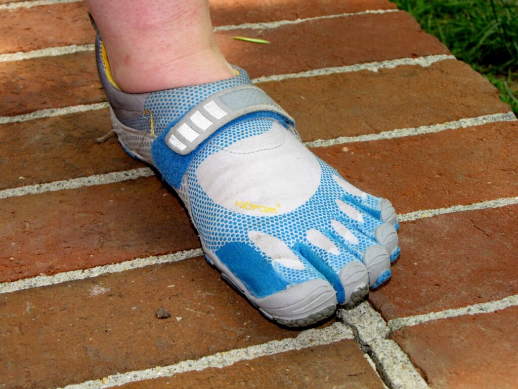 Against a brick floor, a foot wears a blue, gray, and white sneaker whose front makes space for and resembles individual toes.
