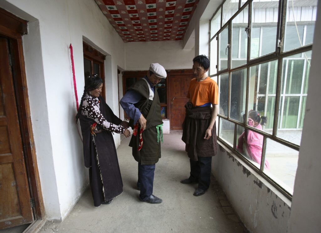 Three people—two in slacks and one in a patterned dress—stand in a hallway with doors on one side of them and a large window on the other. The person in a dress adds a belt to one of the other people’s waists while the third looks on. A child wearing a pink coat peers in at them through the window.