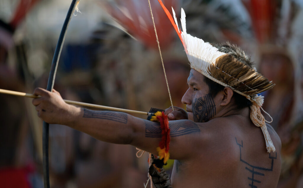 In front of a blurred background of similarly dressed people, a shirtless person with black body markings and a feather headdress draws an arrow on a bow.