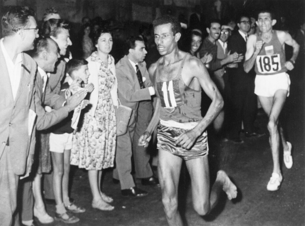 A black-and-white photograph shows a person with the number 11 on their shirt running barefoot in front of others running with sneakers. A crowd lines the race and looks on.