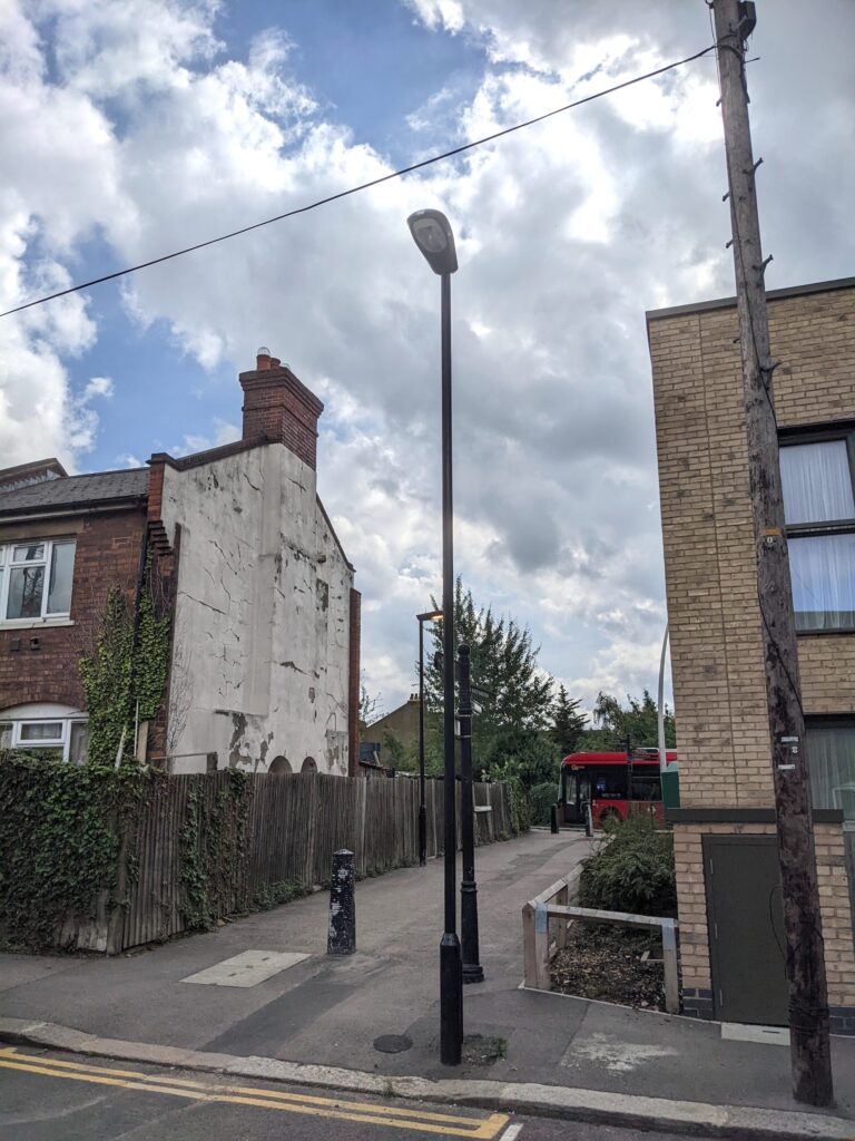 A modern color photograph shows a paved gap between two two-story buildings. A streetlamp is in the foreground, and a red bus is in the background.