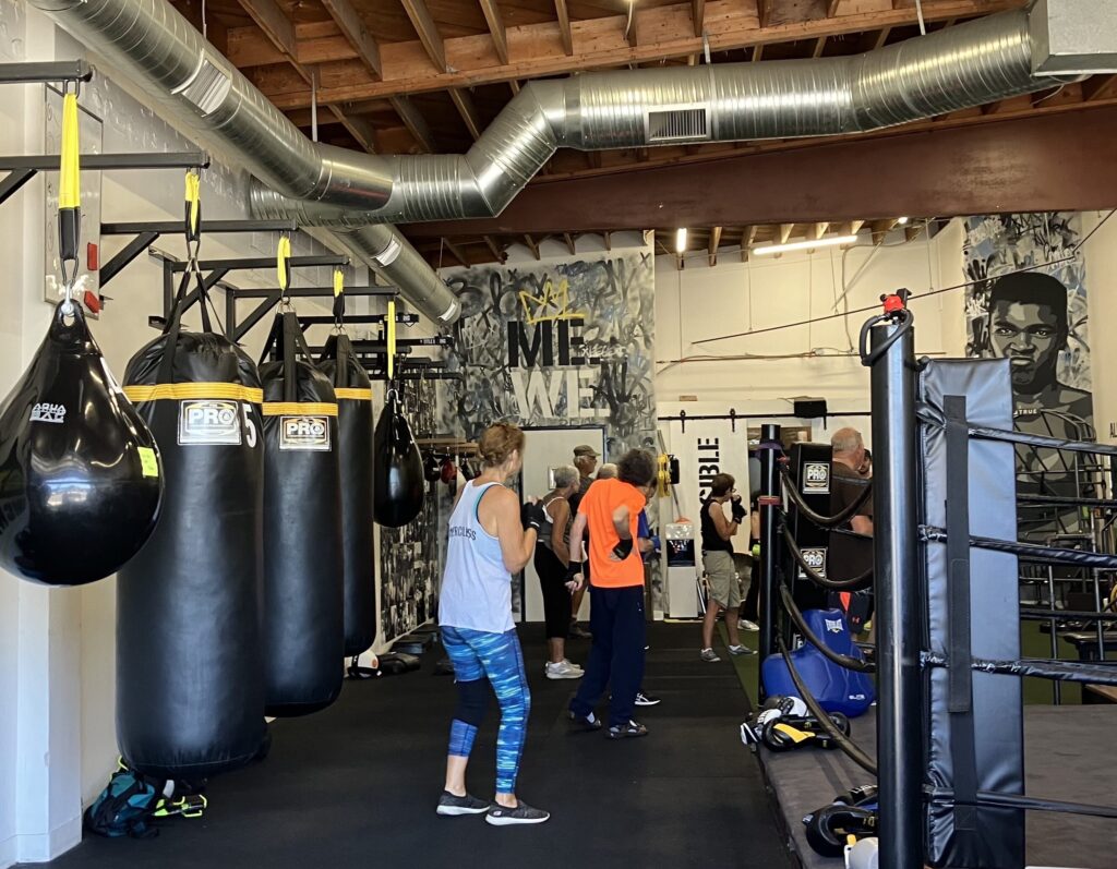 People wearing athletic clothes and open-finger gloves stand on floor mats in a gym near a boxing ring on their right and a wall of punching bags on their left.