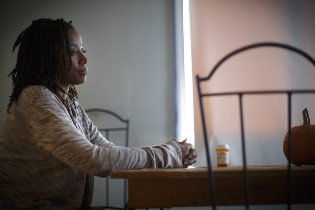 A person with twisted black hair sits at a wooden table and clutches their hands together. A single orange and white pill bottle rests on the table in front of them.