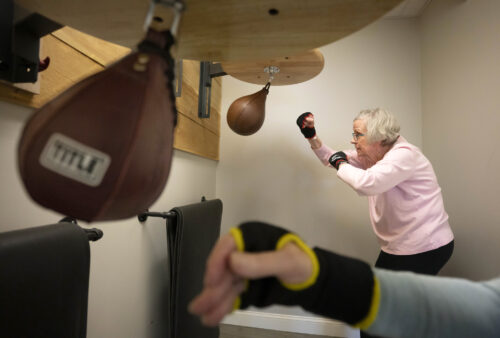 A person with short gray hair wearing a pink, long-sleeved shirt and black pants hits a small swinging punching bag.