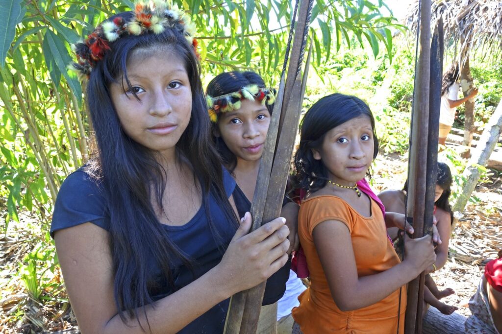 In front of green plants and a few other people, three young people with long black hair wearing feather headpieces hold wooden bows and arrows while looking at the viewer.