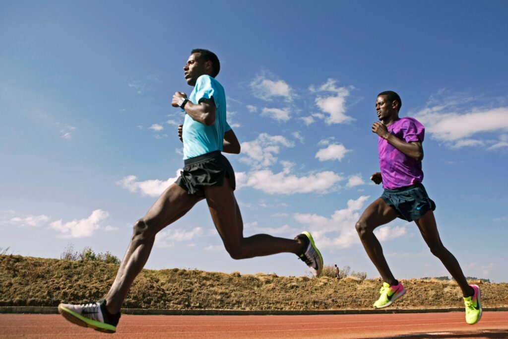 Two people wearing t-shirts, shorts, and sneakers run on an orange track with green shrubs and a clear blue sky in the background.