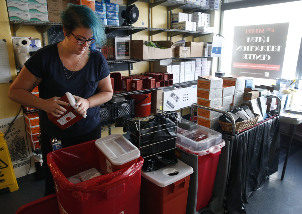 A person with short blue hair wearing black glasses and a navy T-shirt stands by a large, red plastic container and holds a much smaller red and white container. Around them are shelves covered with boxes and other containers, and a table with orange-and-white boxes stacked on it.