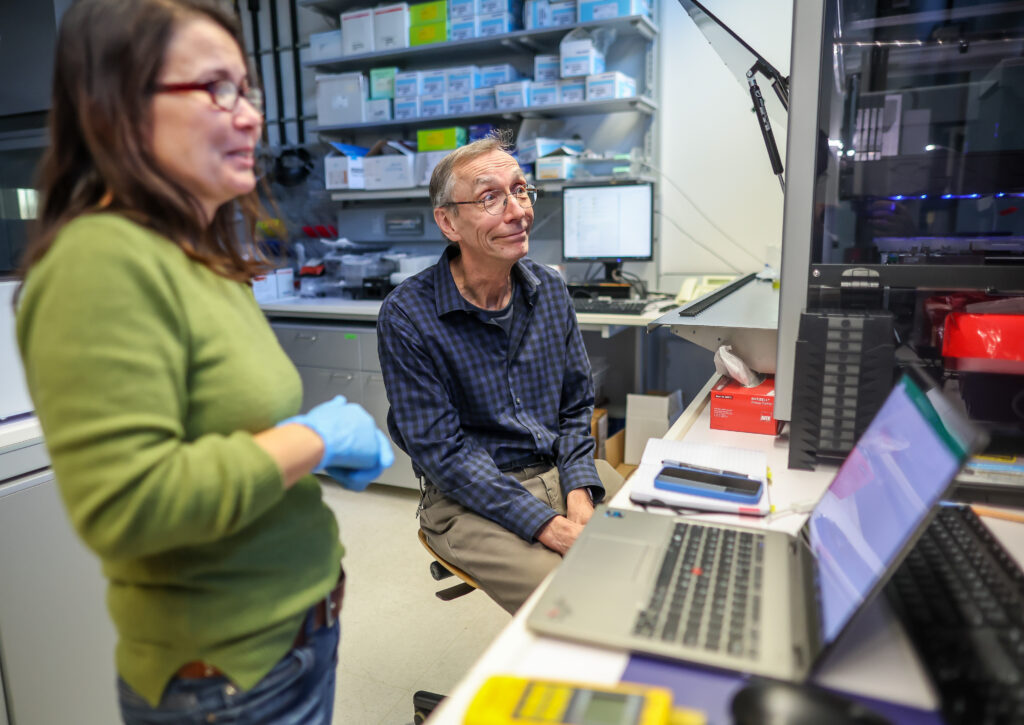 A person with short gray hair wearing glasses, a blue plaid shirt, and khaki pants sits beside a standing person with long brown hair wearing glasses, a green shirt, jeans, and blue medical gloves. They are in a lab that has a desk with a laptop on it, shelves with stacks of white boxes, and a large glass case with a machine in it.
