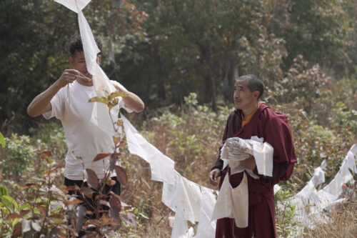 Two people—one in a white T-shirt and the other in burgundy robes—work together to hang a string of white pieces of cloth with black writing on them.