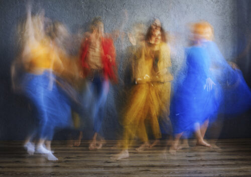 On a wood-paneled floor, four barefoot people wearing different outfits in shades of red, yellow, and blue dance in front of a gray wall. Captured in motion, and therefore blurry, each has several of their dance moves overlapping and visible at once.