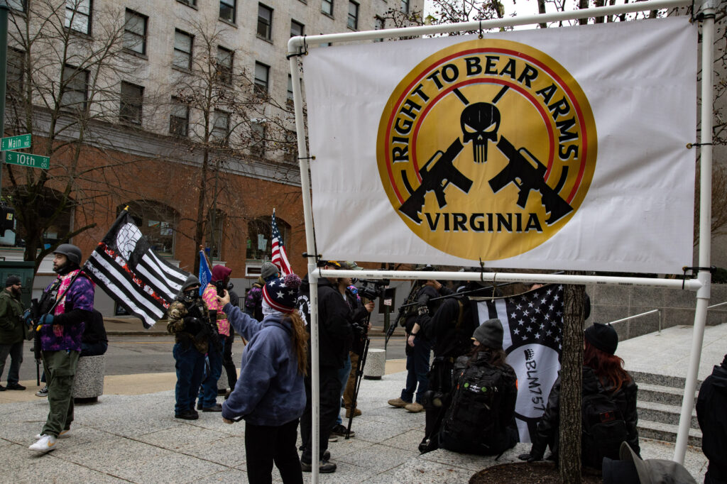 A group of people dressed in black holding flags—some black and white, others red, white, and blue—gather on a sidewalk behind a white banner. The banner features a gold circle with a black graphic of a skull and two guns. Black text reads, “Right to Bear Arms Virginia.”
