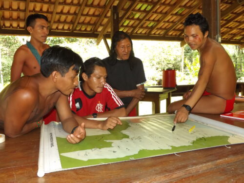 In a wooden structure without walls, three people wearing loincloths and no shirts, and two individuals wearing T-shirts surround and point to spots on a large tan and green map.