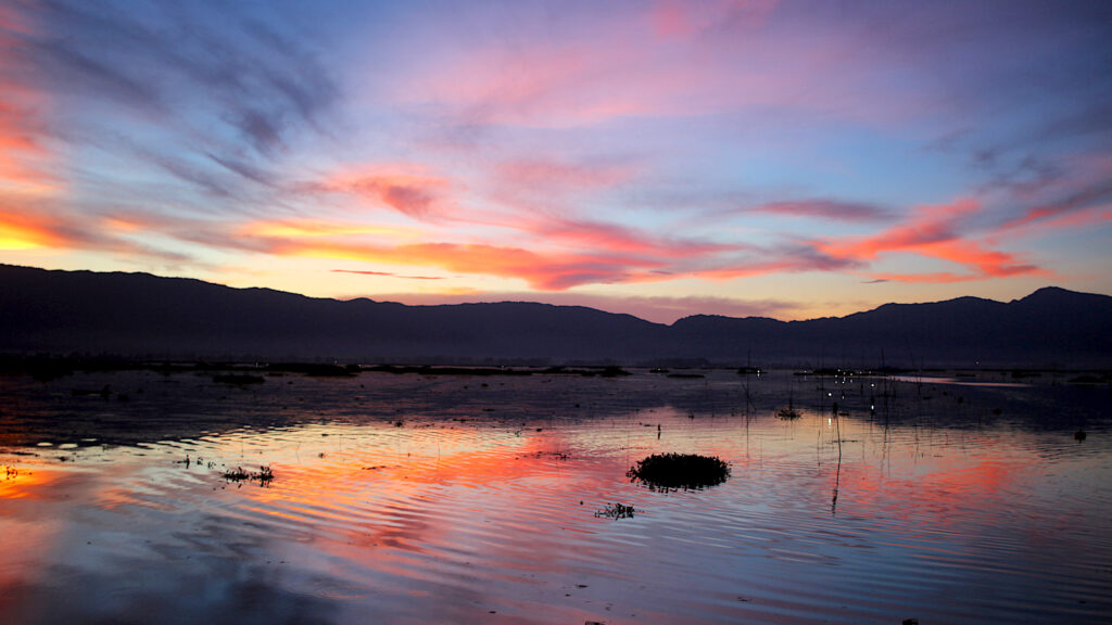 At sunset, a large, gently rippling body of water reflects yellow, orange, and pink light. Small lights scatter along dark silhouetted mountains on the horizon.