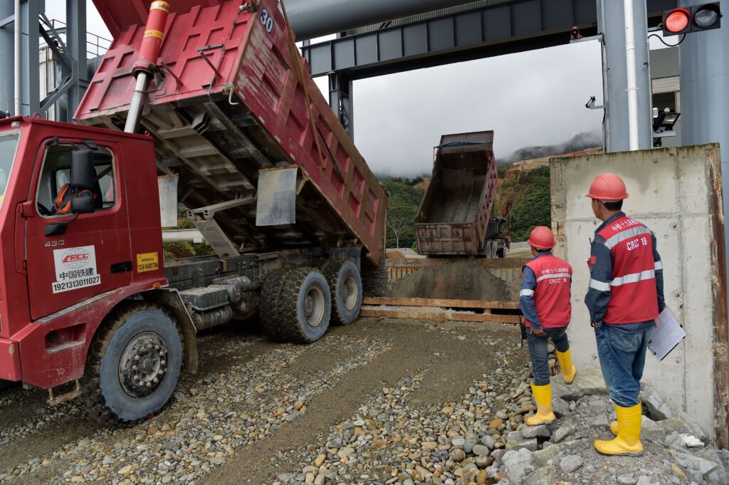 Two people wearing red helmets and vests and yellow boots stand near a large red dump truck by a rocky quarry.
