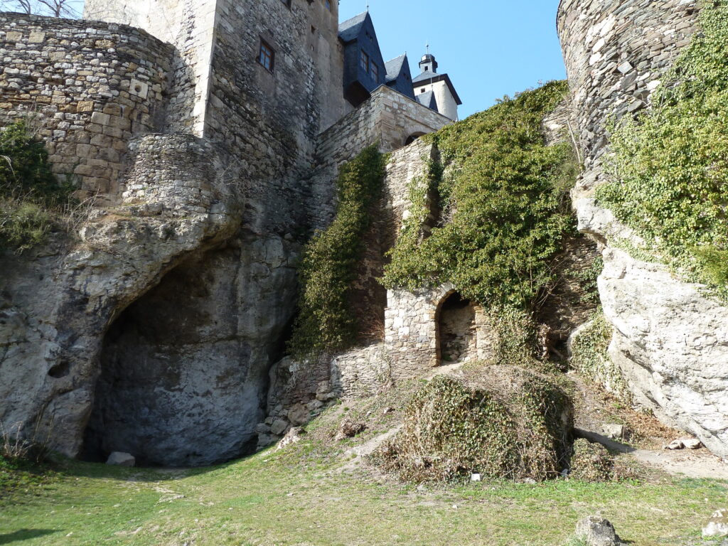 A large, cobblestone building with arches built into its side and covered in green shrubbery towers above a cavernous indent toward the left side of its base.