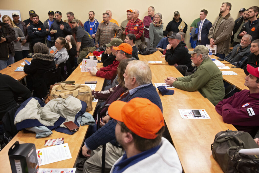 People sit at four long wooden tables arranged in rows. More people stand on the far side of the room, lining a beige wall.