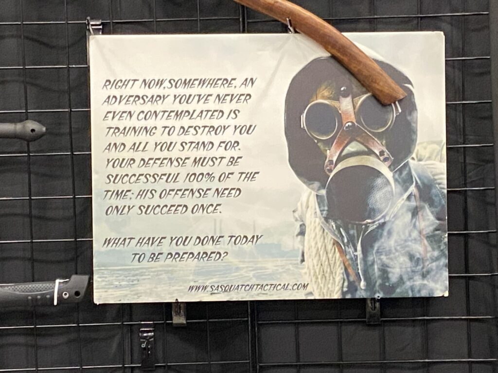 Hung on a wire grid surrounded by knife shafts, a poster features a graphic of a person wearing a black hoodie and gas mask to the right of text that reads, “Right now, somewhere, an adversary you’ve never even contemplated is training to destroy you and all you stand for. Your defense must be successful 100% of the time. His offense need only succeed once. What have you done today to be prepared?”