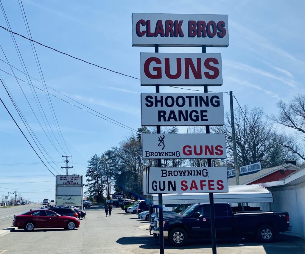 Vehicles are parked outside a white building near a large billboard composed of vertically arranged panels. In red and black text, the signs read, “Clark Bros, Guns, Shooting Range, Browning Guns, Browning Gun Safes.”