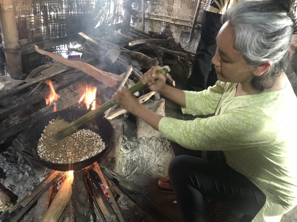 A person with gray hair wearing a green shirt and black pants crouches beside a fire. They use a green tool to stir contents in a black wok that sits on a wood pile over the fire.