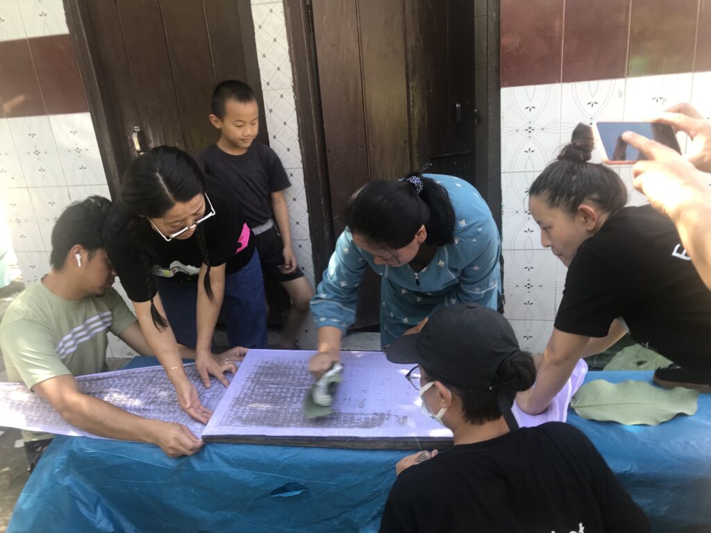 Several people surround and help hold a long piece of white cloth as a person in the center wipes a green object across it, leaving black marks.