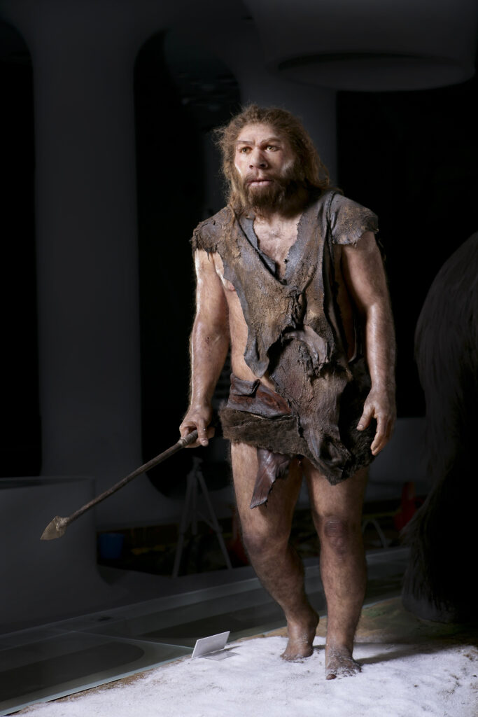 Una figura representa a una persona con piernas peludas, barba y pelo largo, áspero y castaño, que viste ropas marrones rotas y sostiene una lanza.