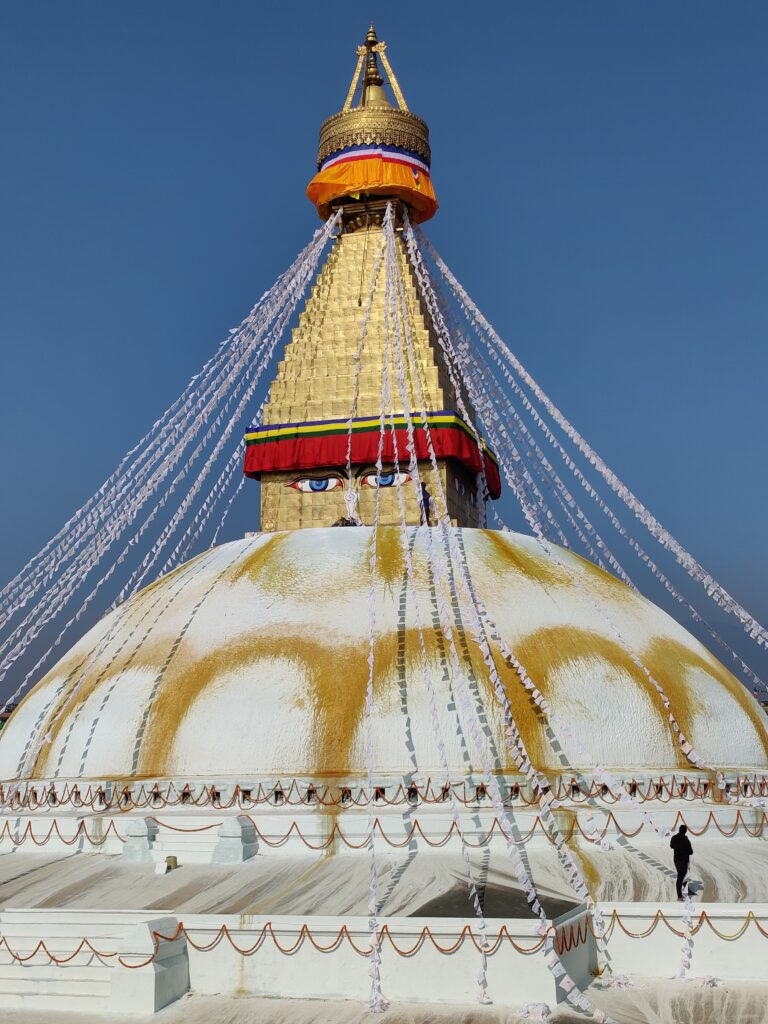 A person stands by a white dome on top of a building. A gold pyramid sits on the dome with blue eyes painted on its base. Strings of white cloths run from the top of the pyramid to the bottom of the dome.