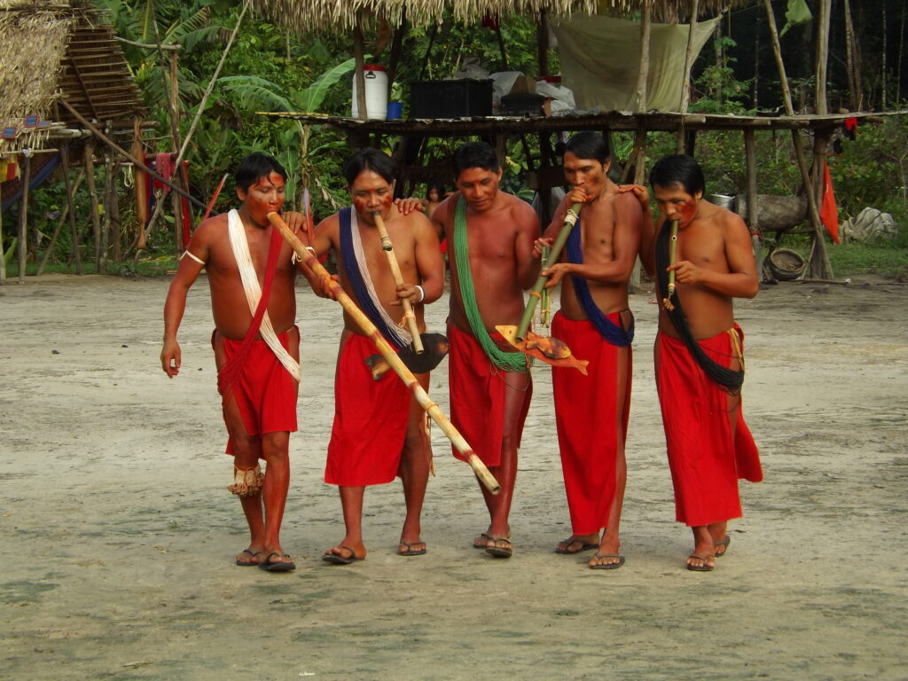 Side by side, a line of five shirtless people wearing long red loincloths and sandals walk while blowing into long bamboo instruments.