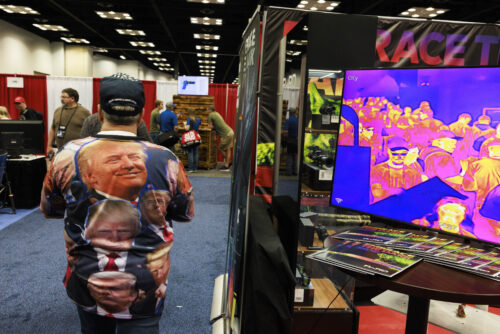 A person wearing a black baseball cap and a shirt covered in a collage of different images of a man’s face walks down a presentation hall. A screen showing many people elsewhere is to their right and a poster with a graphic of a gun hangs over a booth in front.