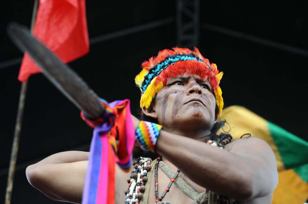 Tirada de um ângulo ascendente, a fotografia mostra uma pessoa sem camisa usando um cocar de penas vermelhas, azuis, amarelas e pretas, uma pulseira colorida e vários colares de contas. Ela está segurando uma bandeira amarela e verde.