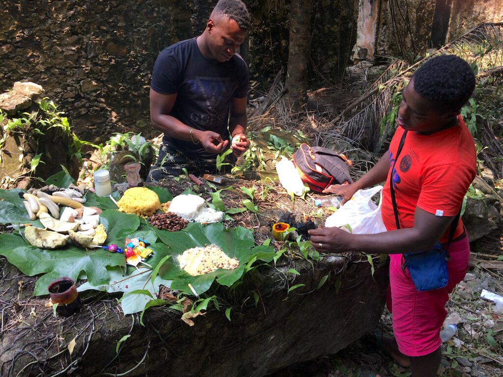 In a forest, two people arrange an array of produce and food on green leaves on an elevated rock. Some of the items include rice, beans, coconuts, and lollipops.
