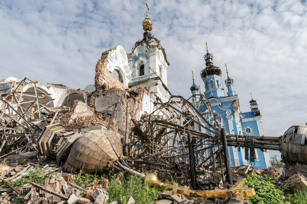 The toppled steeple of a church lies on the ground among other pieces of collapsed metal. A large blue and white building with a gold object on its roof towers in the background.