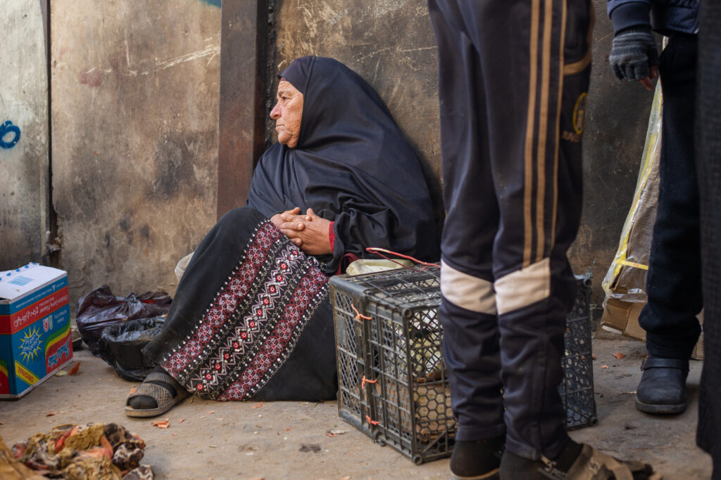 An older person wearing a black headscarf and embroidered skirt sits on the ground, resting against a concrete wall.