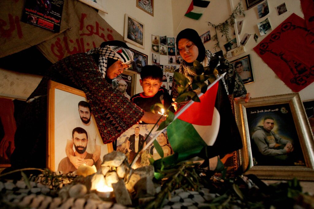 Two adults and a child stand in a room surrounded by lit candles and several framed pictures of people. There is also a green, red, white, and black flag, several white and blue scarves, and pieces of burlap with red text painted on them.