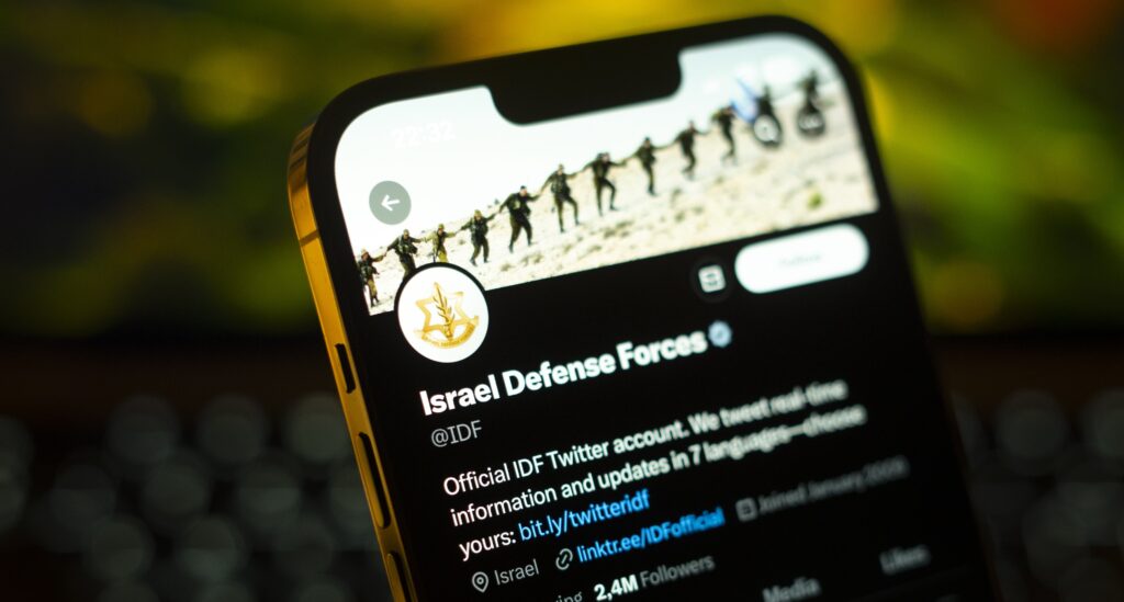 A header image on a cellphone of several people wearing military uniforms and holding hands is above a yellow logo and white text that reads, “Israel Defense Forces.” White text underneath reads, “Official IDF Twitter account. We tweet real-time information and updates in 7 languages—choose yours.”
