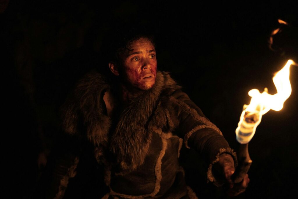 In a pitch-black environment, a person with black smudges on their face wears a fur pelt and holds a lit torch.