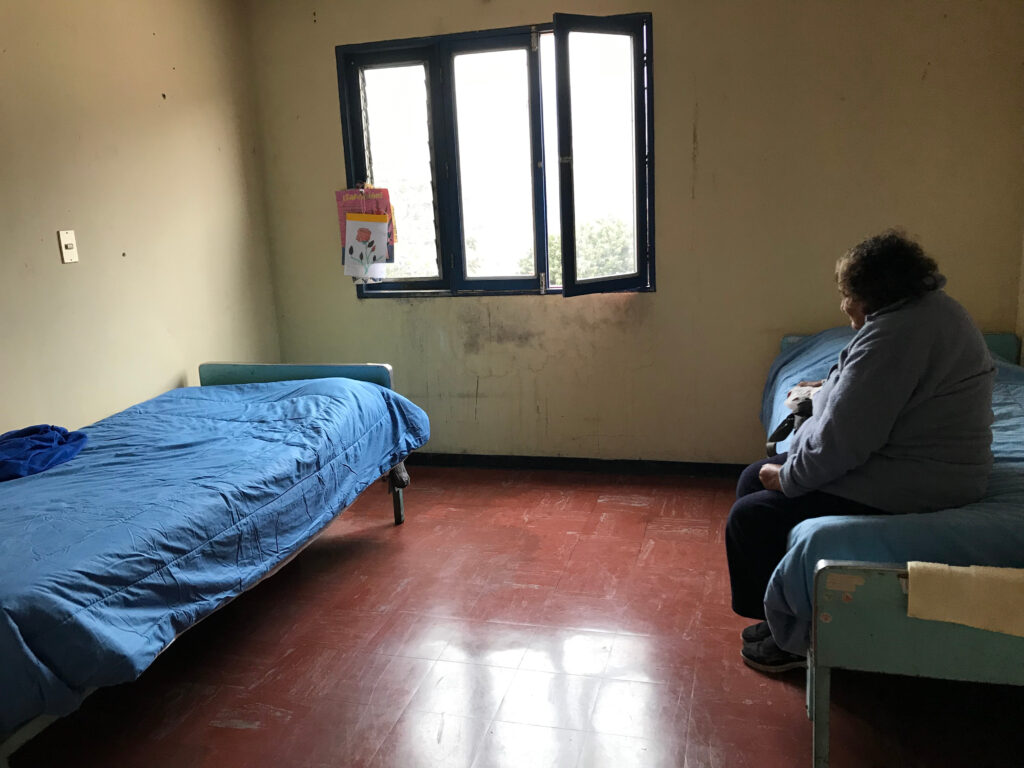 A person with curly gray hair wearing a gray cardigan and slacks sits on a bed covered with a blue comforter and looks across a mostly empty room at a similar bed. An open window is on the back wall.
