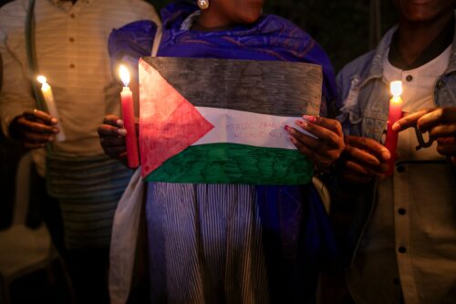 Three people stand in a line holding lit candles. The person in the center holds a piece of paper with a red triangle colored in with marker on the left side, and three horizontal stripes—black, white, and green—on the right. In the white area, text written in red marker reads, “MSJC family in solidarity.”