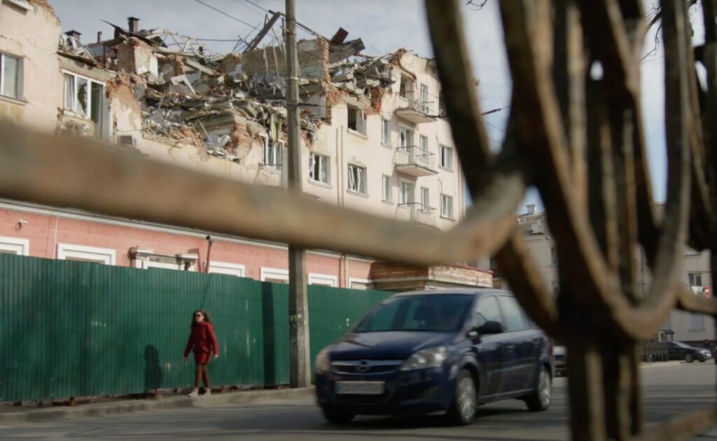 A person wearing a red coat walks down a sidewalk in front of a tall green fence. A black car drives by them, and a building with a collapsed roof is visible on the other side of the fence.
