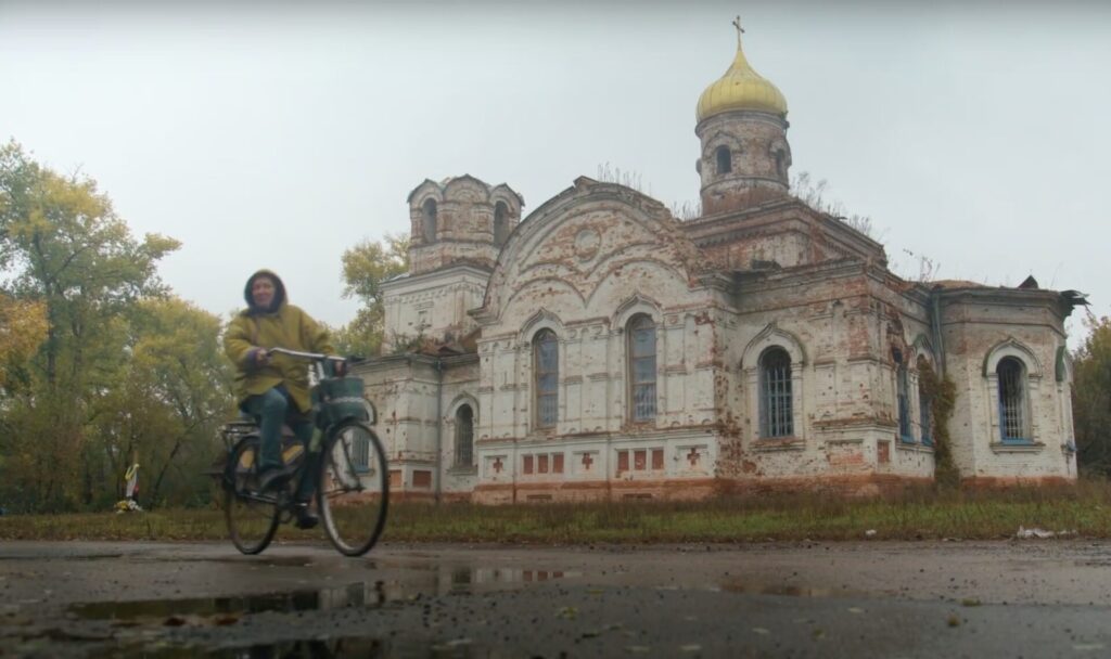 A person rides a bike past a large beige building with a yellow dome and cross on top of it.
