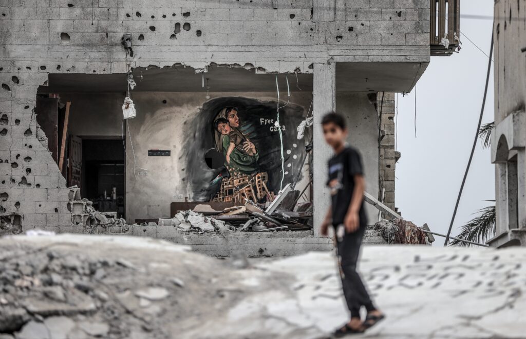 With a child passing in a blurry foreground, a mural depicting a person with a headscarf carrying a child to the left of the words “Free Gaza” covers a wall inside a bombed brick building.