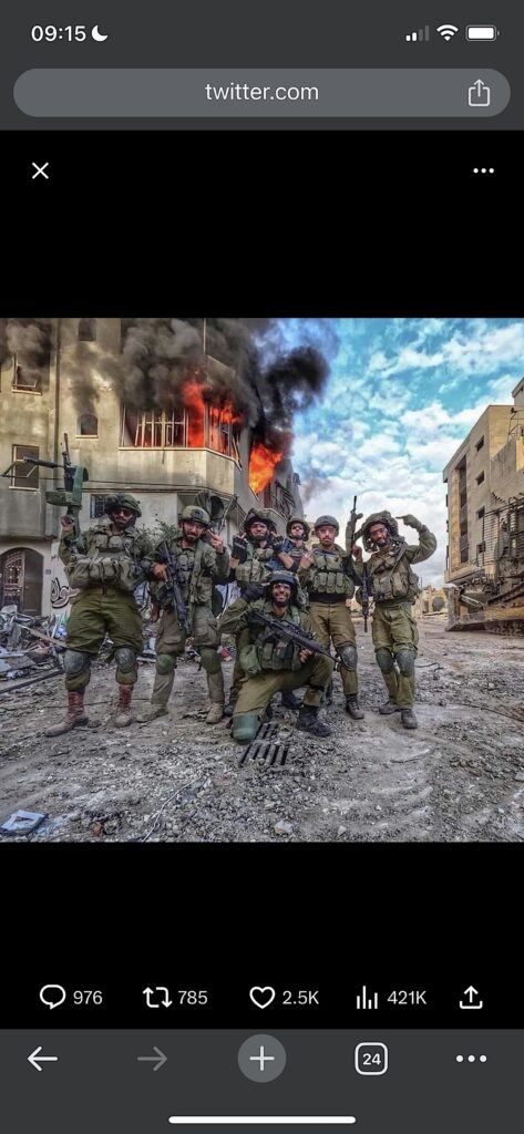 A group of people in green camo uniforms and helmets hold large guns and pose in front of a burning building surrounded by other destroyed buildings and debris.