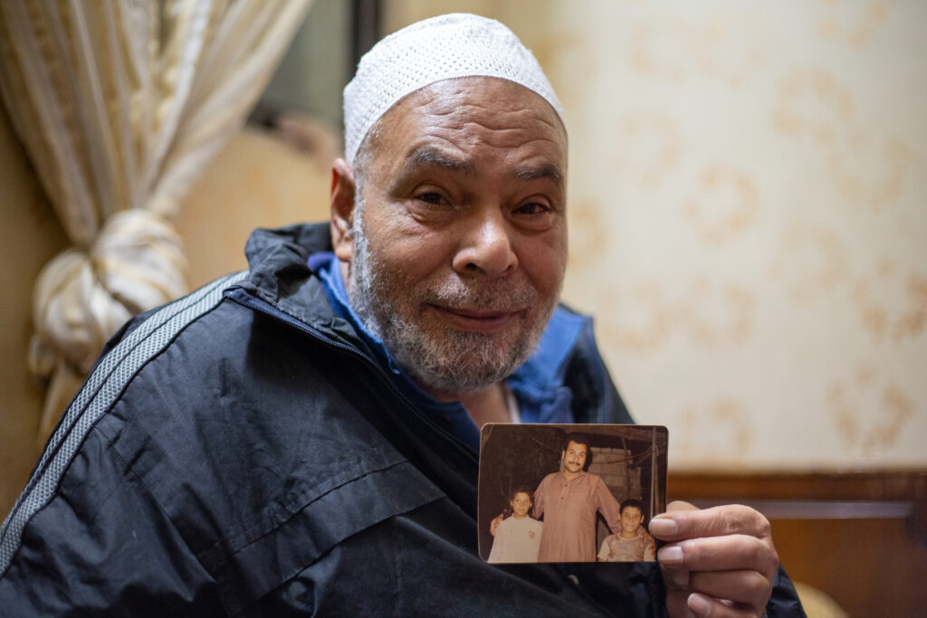 An older man wearing a white kufi cap and navy-blue windbreaker holds up a photo of a younger man with two children.