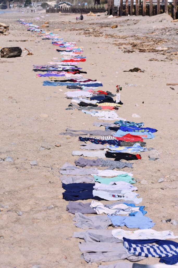 Several pieces of children’s clothing lie in a long line down the length of a sandy shore.