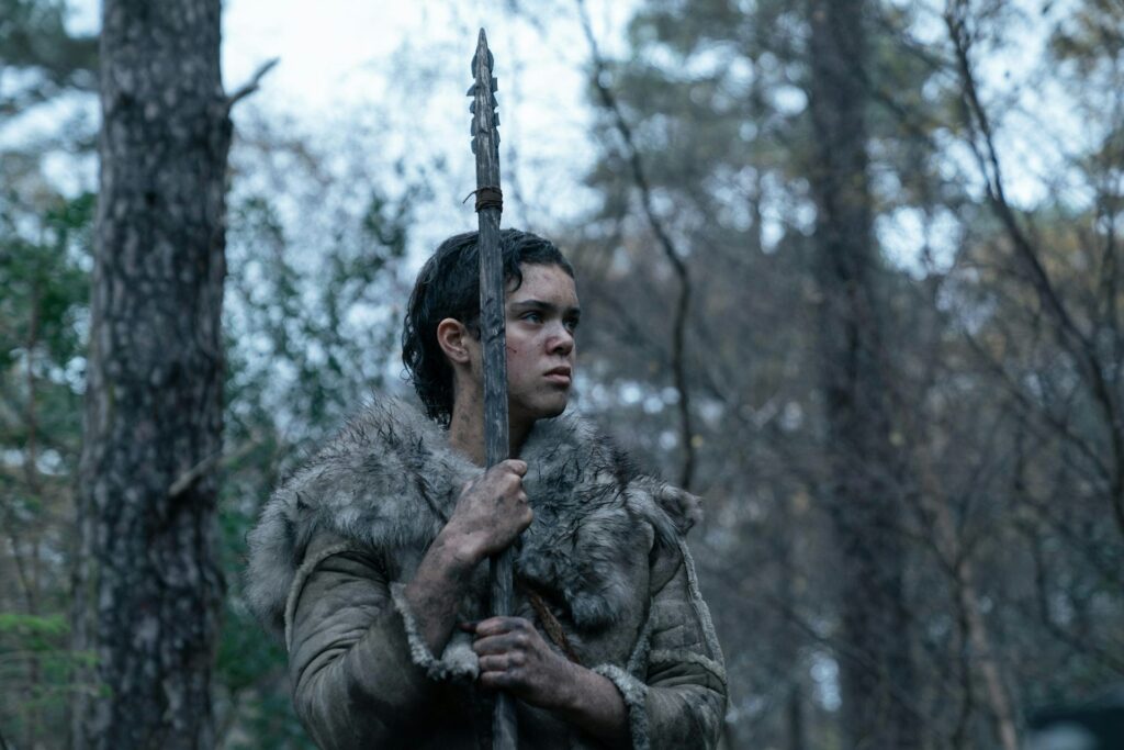 In a forest of leafless trees, a person wearing a fur pelt holds a spear vertically.